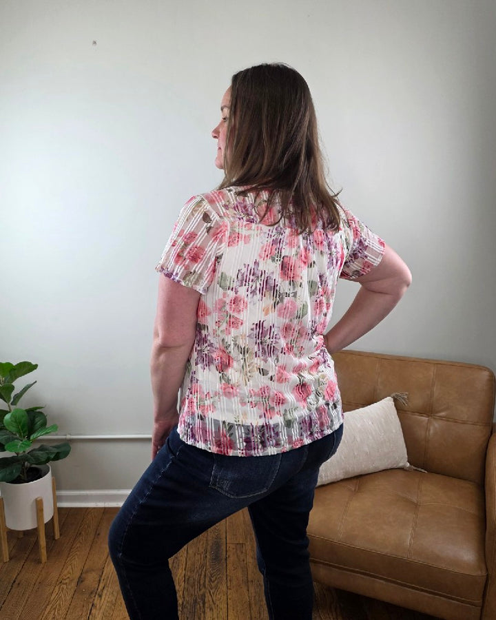 A woman with straight brown hair smiles indoors, wearing the Miranda Floral Print, Flutter Sleeve Blouse and jeans from Cosmos + Clover Boutique. She stands with one hand on her hip in front of a tan couch, pillow, green potted plant, and light-colored wall.