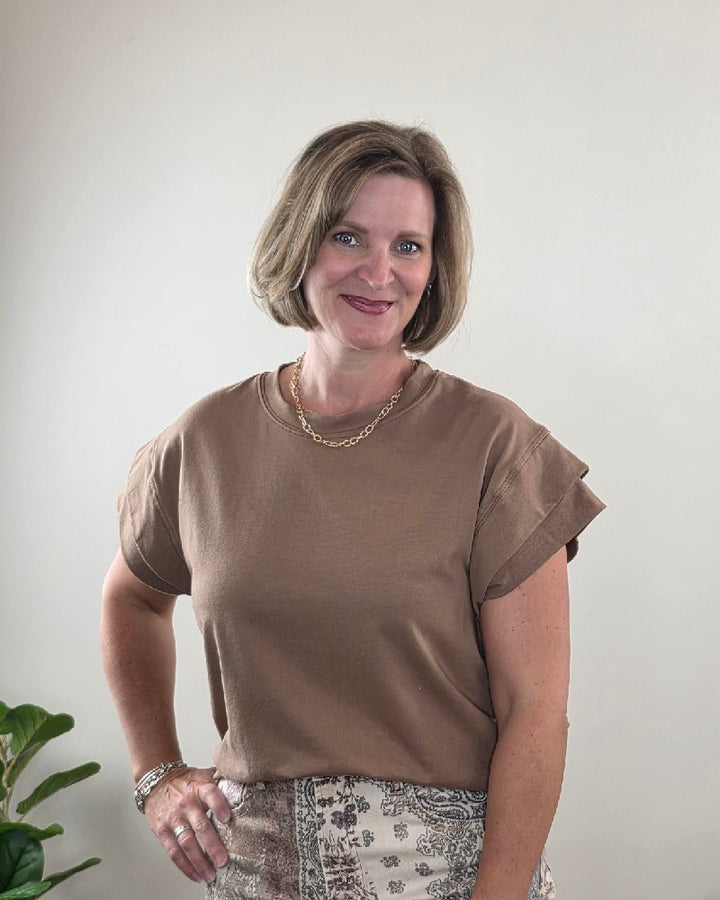 A woman with short blond hair, wearing the Les Amis Neriah Short Sleeve Mocha Top with Layered Sleeve and a patterned skirt, smiles while standing in front of a plain light-colored wall. A green plant is partially visible in the lower left corner.
