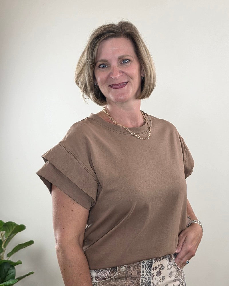 A woman with short blonde hair wears the Les Amis Neriah Short Sleeve Mocha Top with Layered Sleeve and a patterned skirt, smiling at the camera. She stands in front of a plain light background, with a leafy plant partially visible on the left.