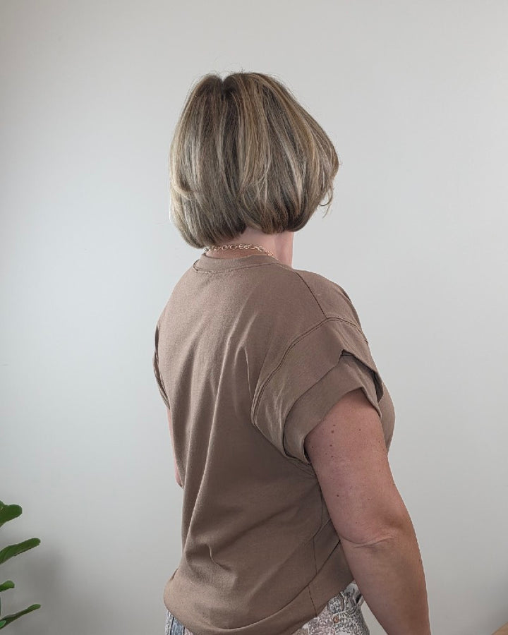 A woman with short blonde hair stands facing away from the camera against a plain light gray wall, wearing the Les Amis Neriah Short Sleeve Mocha Top with Layered Sleeve and patterned pants. Part of a green plant is visible in the lower left corner.