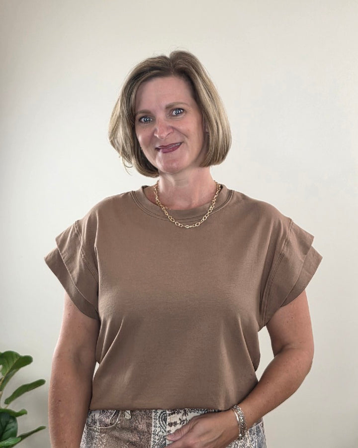 A woman with short blonde hair, dressed in the Les Amis Neriah Short Sleeve Mocha Top with Layered Sleeve styled for work and accessorized with a gold chain necklace, stands in front of a light wall. A green plant is partially visible in the lower left corner.