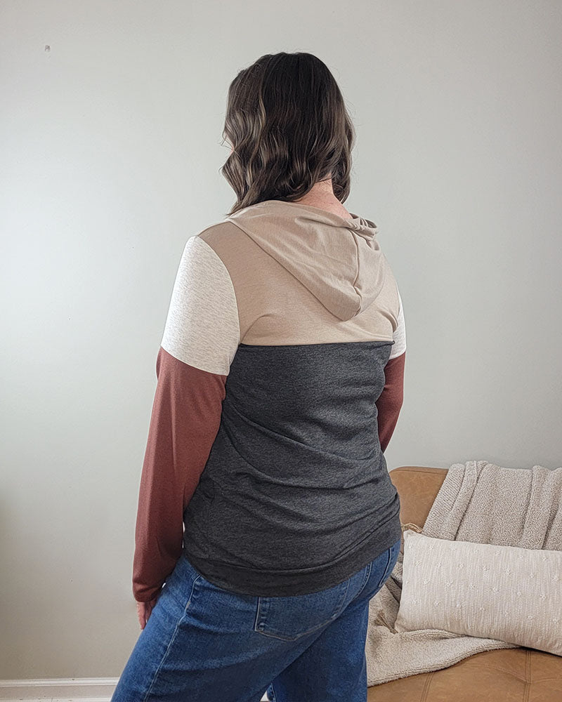 A person with wavy brown hair, wearing the Heimish Jamie Color Block Hoodie in beige, gray, and rust plus blue jeans, stands with their back to the camera in a neutral room with a tan couch and textured pillow.