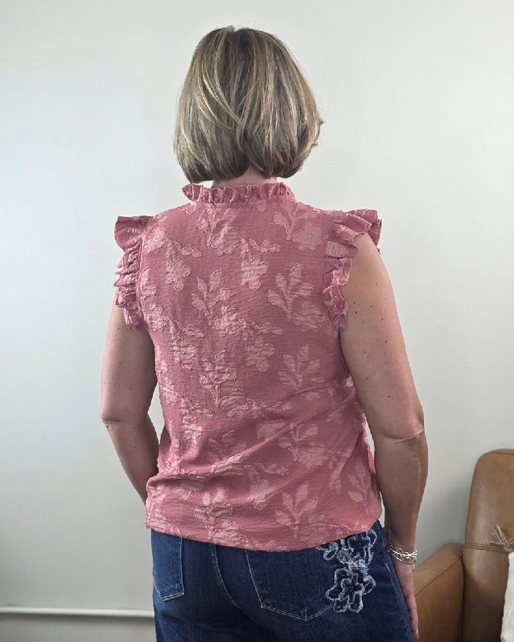 A woman with short blonde hair wears the Nicole Ruffle Edge Top by Heimish, a dusty mauve blouse featuring a ruffle flower pattern and a flowy, flattering fit. She pairs it with blue jeans decorated with floral embroidery and stands with her back to the camera.