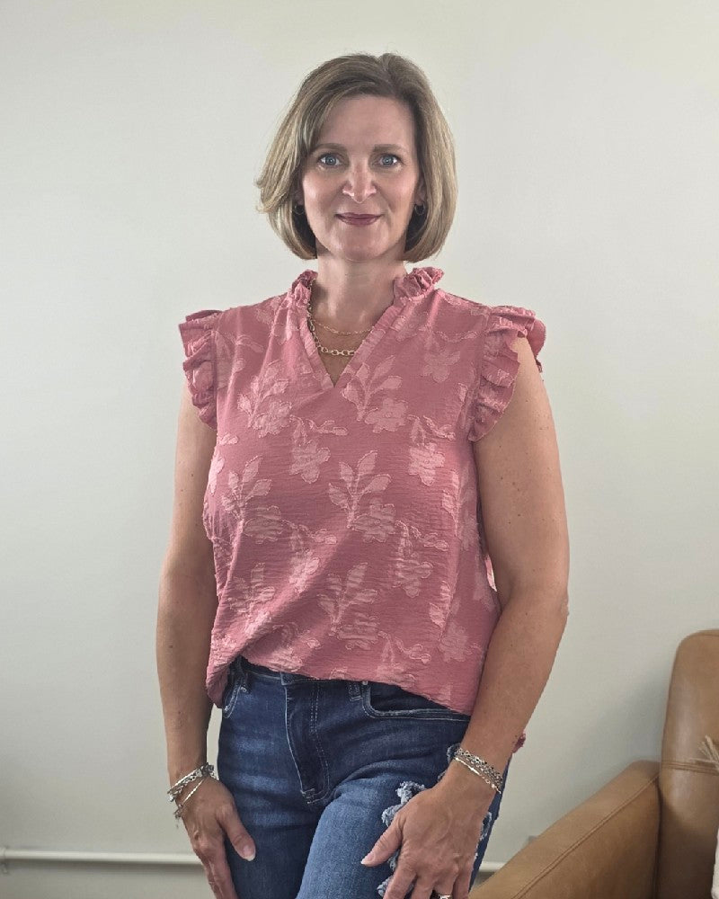A woman with short blonde hair models the Nicole Ruffle Edge Top by Heimish, a dusty mauve blouse adorned with a ruffle flower pattern, paired with blue jeans. She stands indoors beside a tan chair, smiling at the camera against a plain, light-colored wall. The top’s flowy and flattering fit lends an elegant touch to her ensemble.