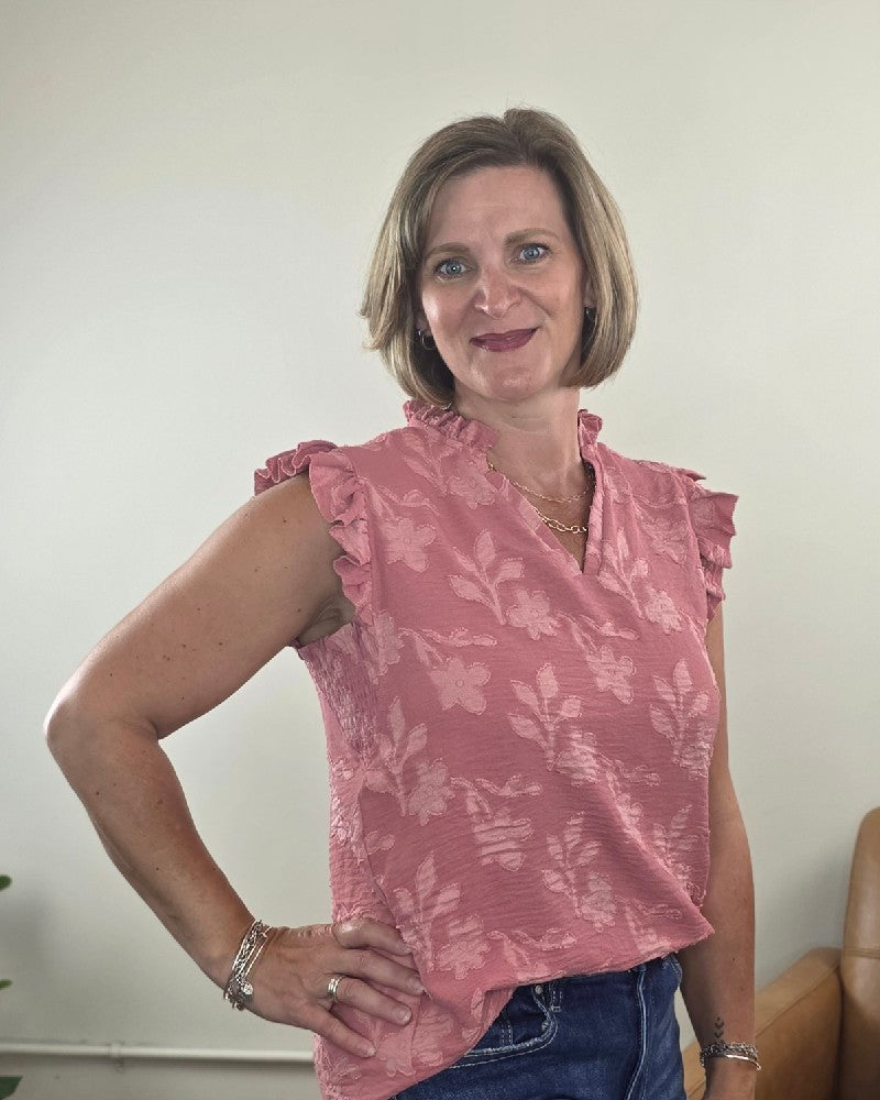 A woman with short light brown hair is smiling, wearing the Heimish Nicole Ruffle Edge Top in dusty mauve paired with blue jeans, standing indoors against a light-colored wall.