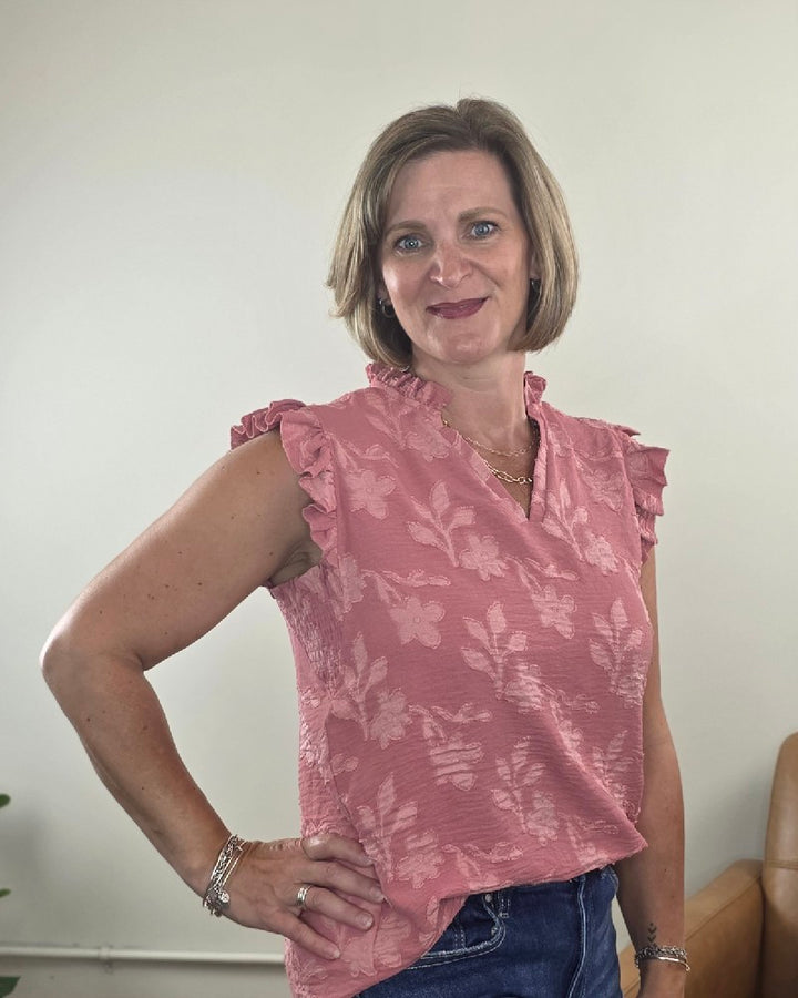 A woman with short light brown hair is smiling, wearing the Heimish Nicole Ruffle Edge Top in dusty mauve paired with blue jeans, standing indoors against a light-colored wall.