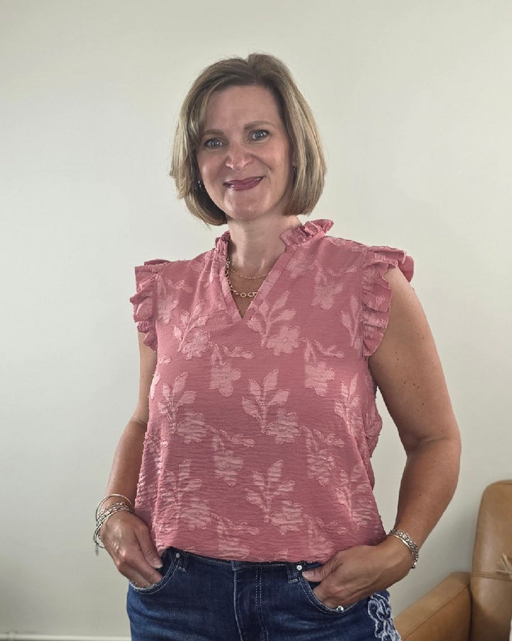 A woman with short blonde hair is smiling, wearing the Heimish Nicole Ruffle Edge Top and blue jeans. She stands indoors against a plain wall, with a tan chair partially visible on the right.