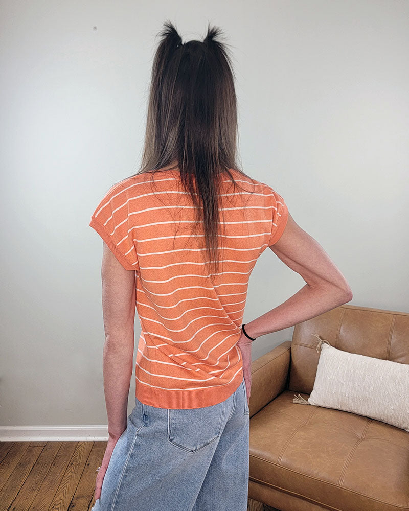 A person with long brown hair in half-up pigtails stands facing away, wearing the Staccato Hallie Striped Short Sleeve Sweater in orange and white with light blue jeans, next to a tan sofa and a white pillow.