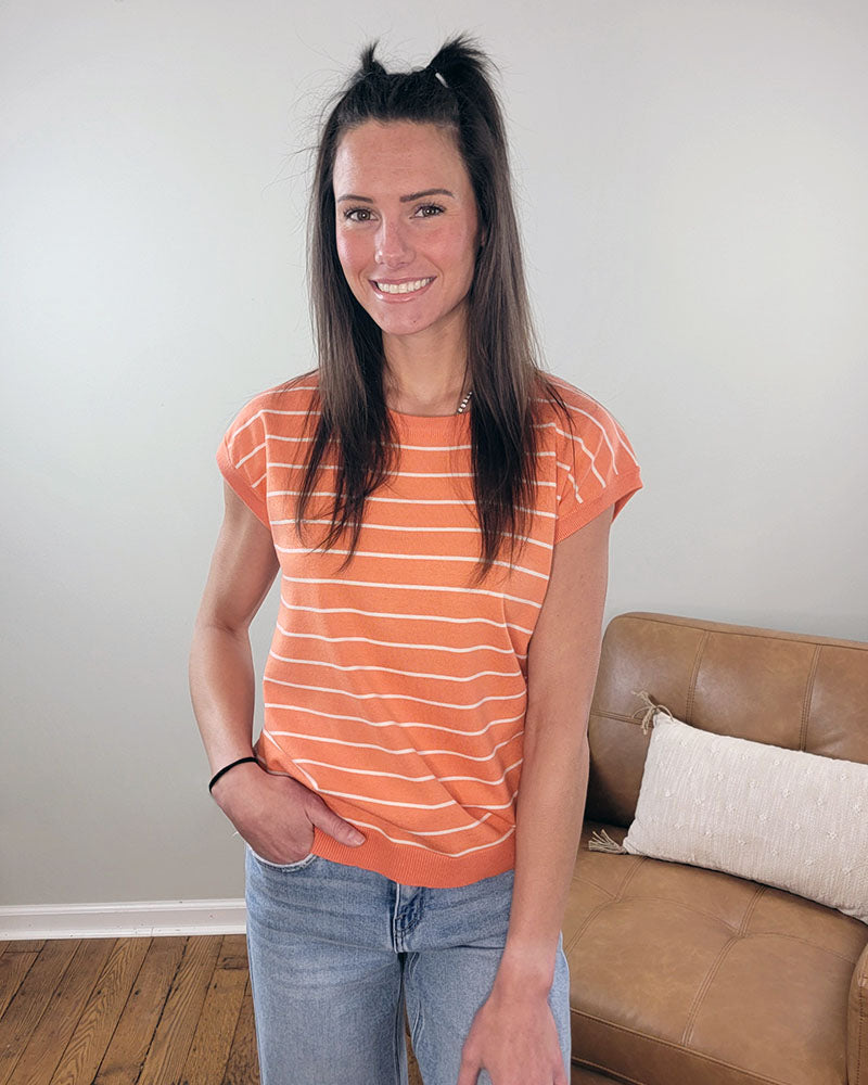 A woman with long brown hair in half-up ponytails wears the Staccato Hallie Striped Short Sleeve Sweater in orange and white, paired with blue jeans, smiling in a living room with a tan couch and white pillow behind her.
