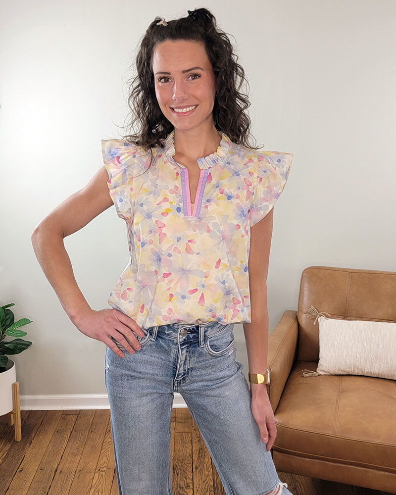 A woman with curly hair smiles indoors, wearing the Staccato Paxton Floral Blouse with ruffle sleeves and light blue jeans. A tan couch and potted plant appear in the background.