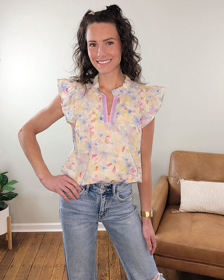 A woman with curly hair smiles indoors, wearing the Staccato Paxton Floral Blouse with ruffle sleeves and light blue jeans. A tan couch and potted plant appear in the background.