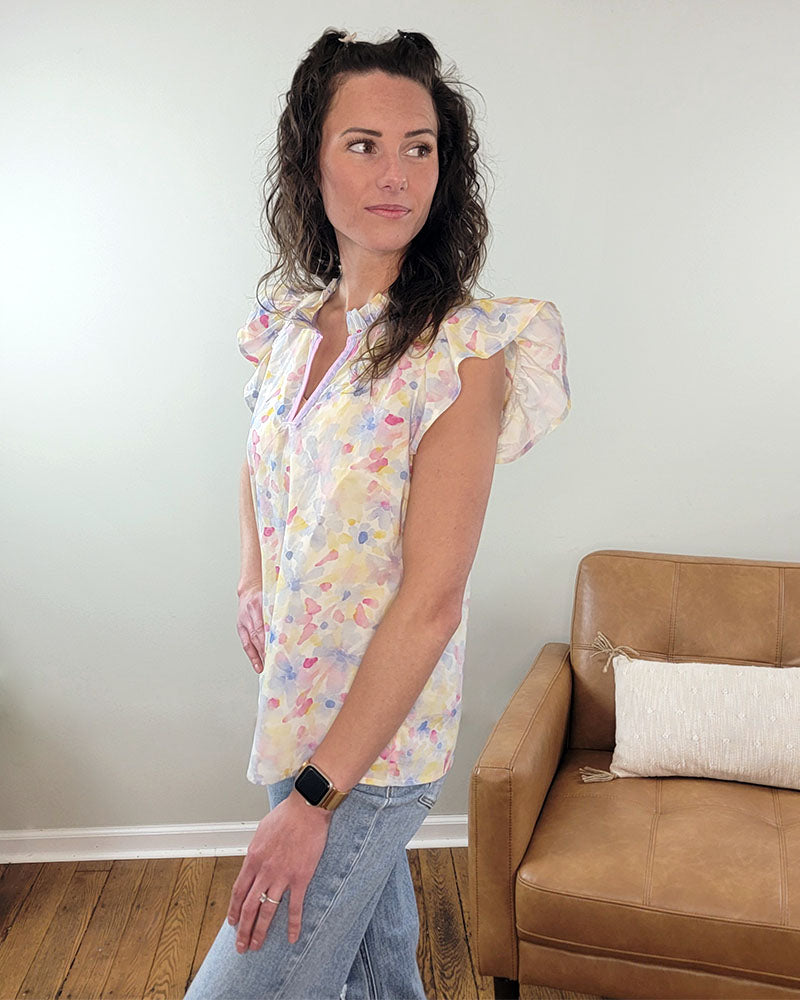 A woman with wavy brown hair wears the Staccato Paxton Floral Blouse with ruffle sleeves and light blue jeans, standing indoors next to a tan armchair and looking to the side.