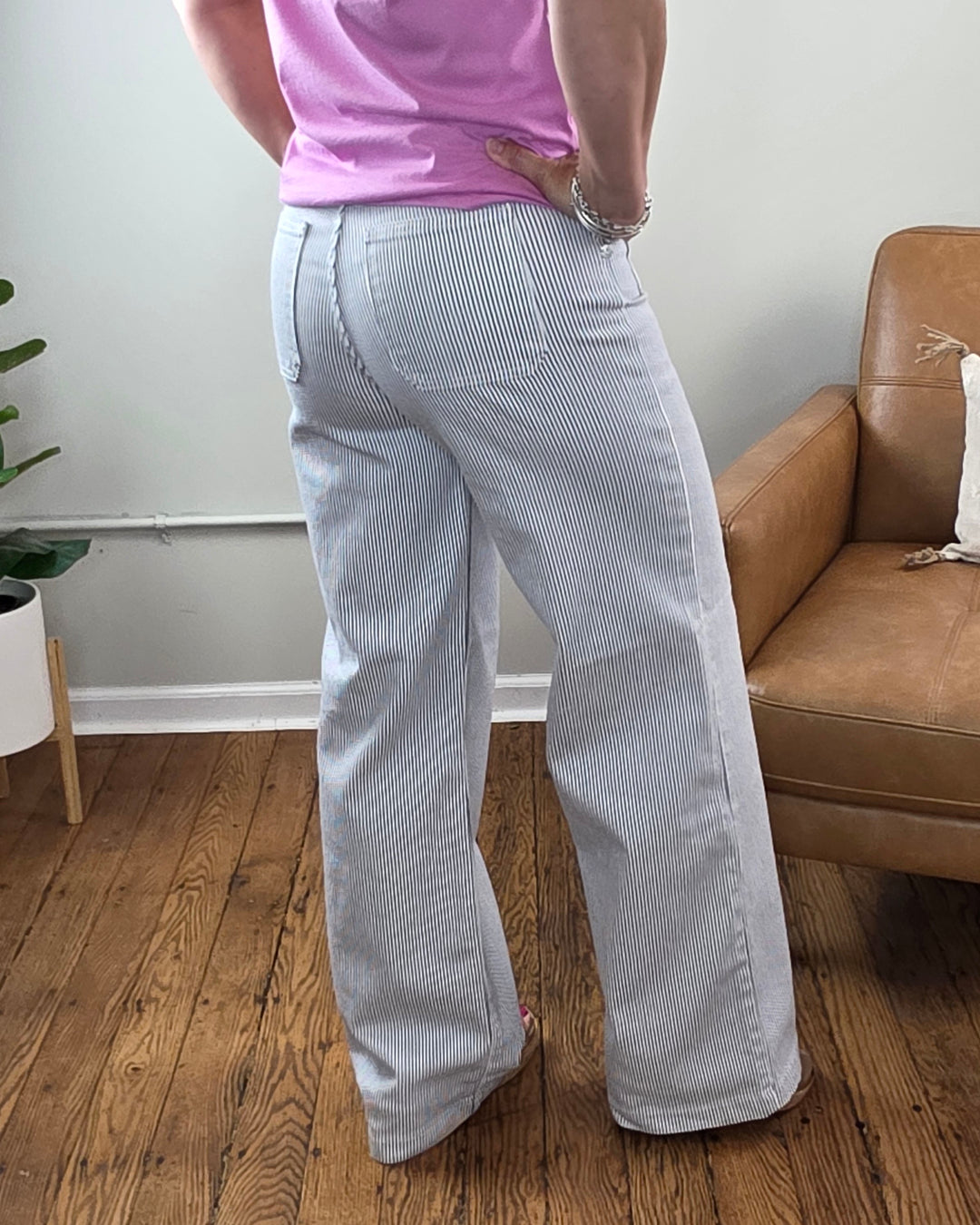 Wearing the Payten Stripe Wide Leg Jeans from JUST USA in light blue and white pinstripes, paired with a pink shirt, this person stands on a wooden floor next to a tan couch and a potted plant.