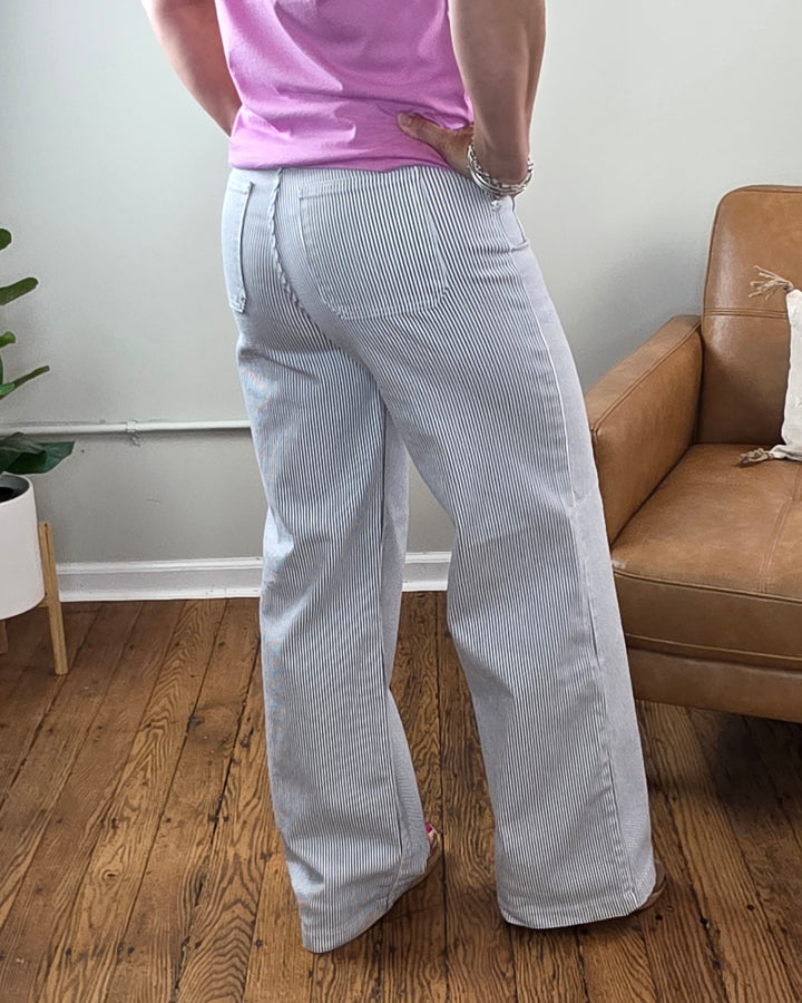 Wearing the Payten Stripe Wide Leg Jeans from JUST USA in light blue and white pinstripes, paired with a pink shirt, this person stands on a wooden floor next to a tan couch and a potted plant.