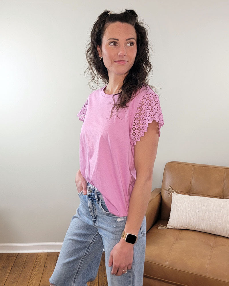 A woman with wavy brown hair wears the Staccato Beverly Top— a pink 100% cotton crew neck with scalloped lace sleeves—paired with light blue jeans and a smartwatch, standing indoors by a tan couch and softly smiling.