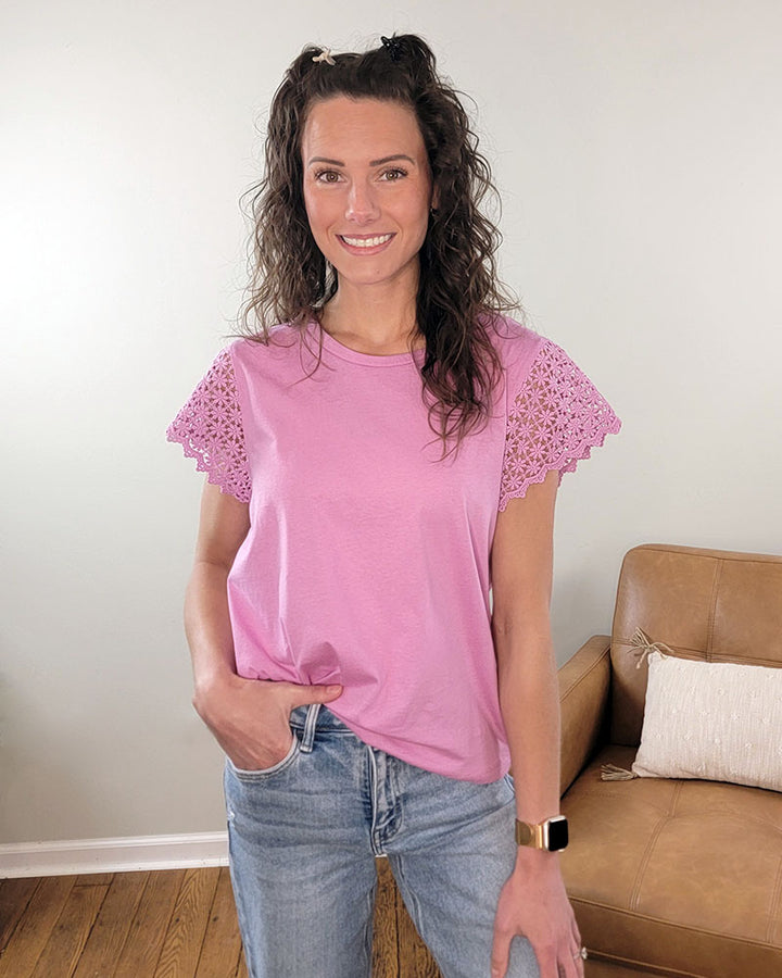 A woman with curly brown hair smiles indoors, wearing the Staccato Beverly Top—a light pink, 100% cotton crew neck with scalloped lace sleeves—and blue jeans. She stands by a beige sofa with a white pillow, one hand in her pocket.