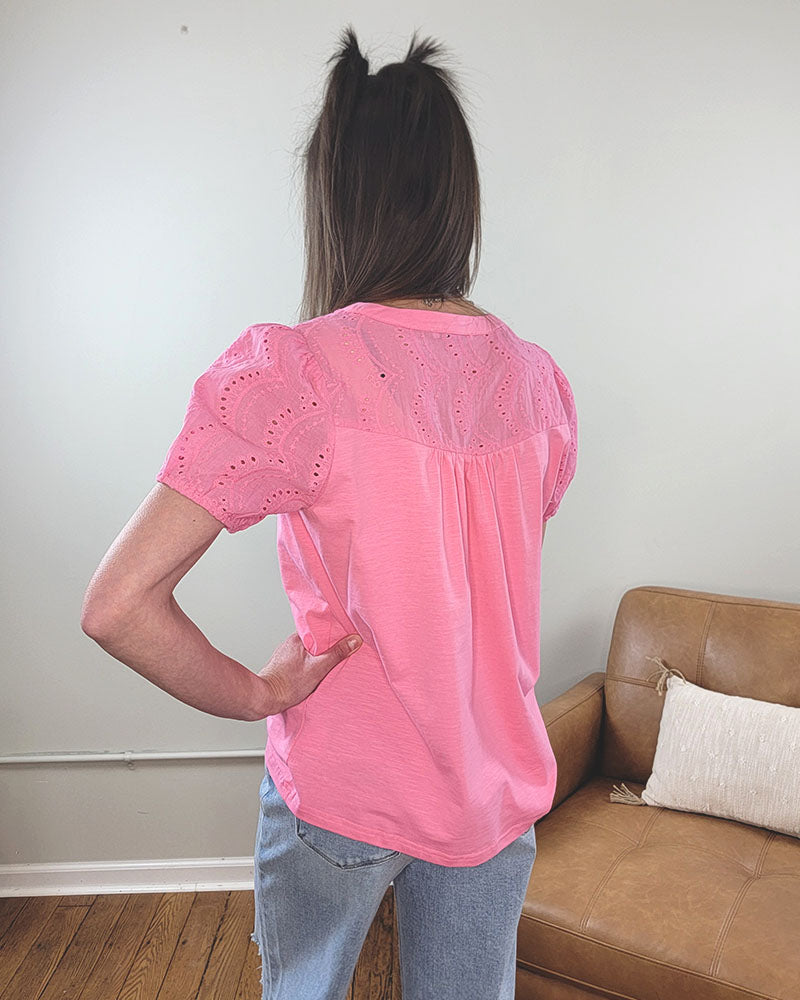 A woman with long brown hair stands with her back to the camera, wearing the Staccato Everlee Blouse in fuchsia with eyelet puff sleeves and light blue jeans, in a room featuring a brown sofa and a cushion.