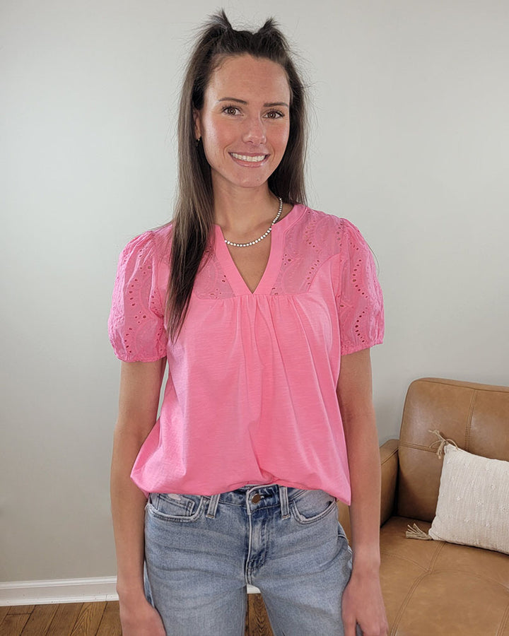 A woman with long brown hair, wearing the Staccato Everlee Blouse in fuchsia and blue jeans, stands smiling in a living room with a tan couch and white pillow in the background.