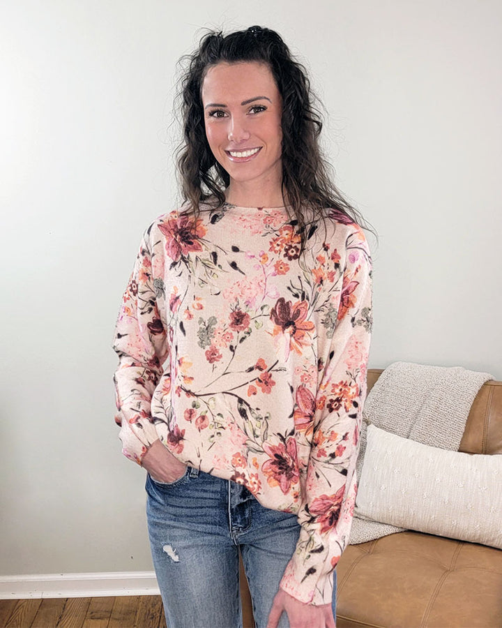 A woman with long wavy hair smiles indoors in front of a light wall and brown couch, wearing the Fate Selma Floral Sweater—a light crewneck with watercolor florals—paired with blue jeans.