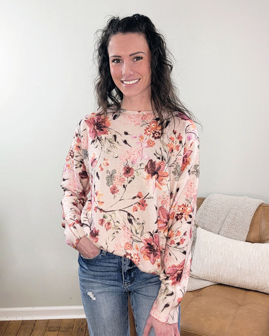 A woman with long wavy hair smiles indoors in front of a light wall and brown couch, wearing the Fate Selma Floral Sweater—a light crewneck with watercolor florals—paired with blue jeans.