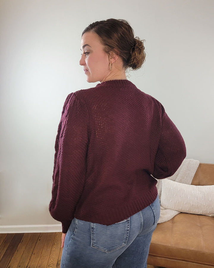 A woman with brown hair in a bun stands indoors, looking over her shoulder while wearing the Doe &amp; Rae Peyton Knit Sweater with Puff Sleeve in burgundy and blue jeans. A tan couch topped with a white blanket appears in the background.
