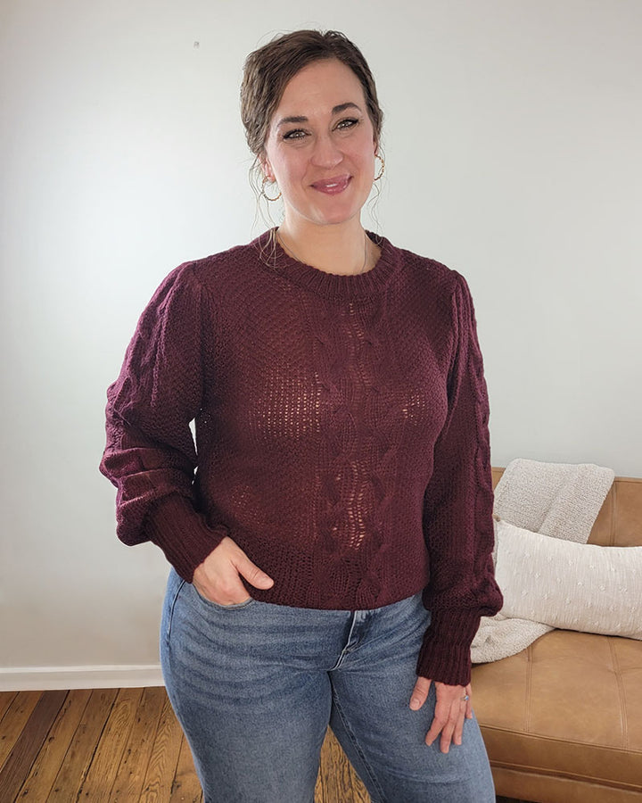 A woman with brown hair smiles indoors, wearing the Doe &amp; Rae Peyton Knit Sweater with Puff Sleeve in burgundy and blue jeans. Behind her is a tan couch with a light blanket on a wooden floor.