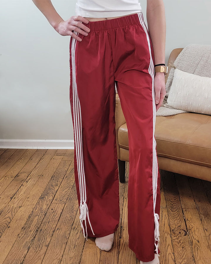 A person wears Love Poem’s Taylor Striped Bow Pants—retro red with white stripes—standing on a wooden floor by a tan sofa, only their lower half visible.