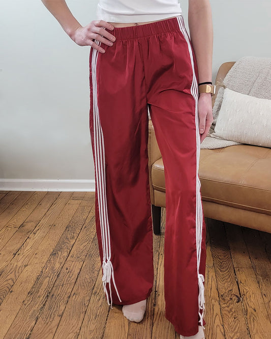 A person wears Love Poem’s Taylor Striped Bow Pants—retro red with white stripes—standing on a wooden floor by a tan sofa, only their lower half visible.