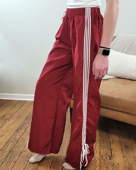A person in Love Poem’s Taylor Striped Bow Pants, paired with a white top and Apple Watch, stands on wooden flooring next to a tan couch with a white pillow—showcasing timeless retro style.