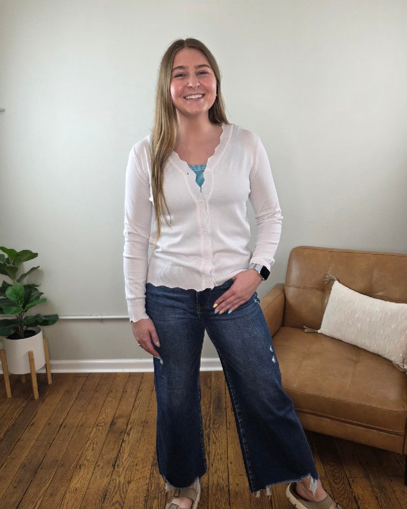 A woman with long light brown hair smiles indoors, wearing the Lovesoft Romi Scalloped Edge Cardigan over a blue top, wide-leg jeans, and sandals. Behind her is a tan couch with a pillow and a potted plant on a wooden floor.