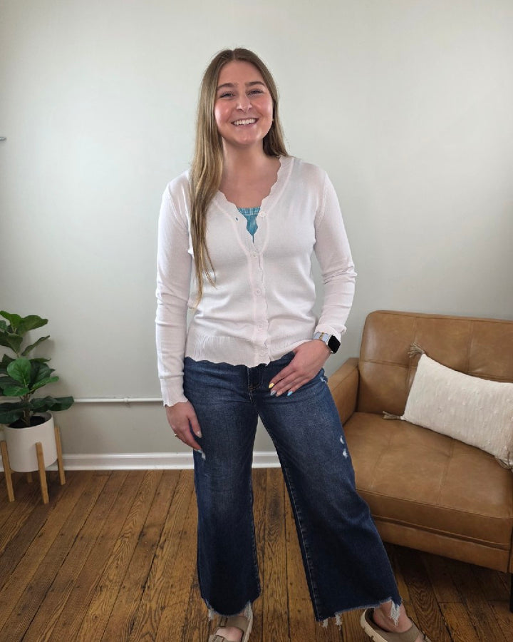 A woman with long light brown hair smiles indoors, wearing the Lovesoft Romi Scalloped Edge Cardigan over a blue top, wide-leg jeans, and sandals. Behind her is a tan couch with a pillow and a potted plant on a wooden floor.