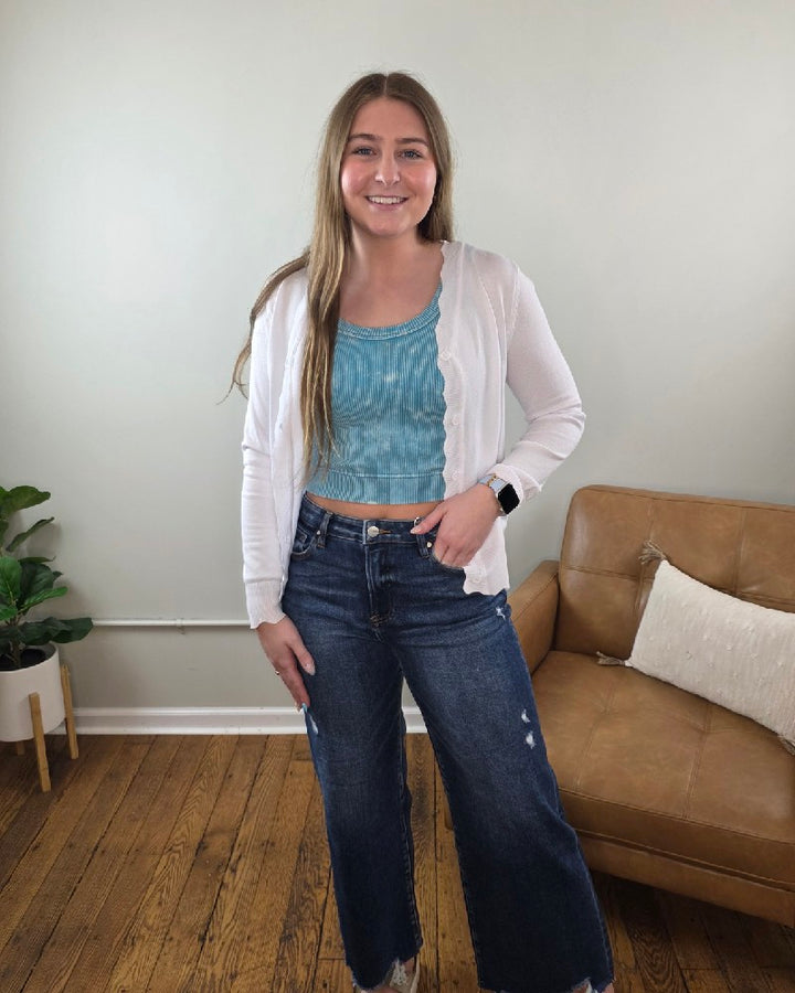 A young woman with long brown hair smiles indoors, wearing the Lovesoft Romi Scalloped Edge Cardigan over a blue top, wide-leg jeans, and a smartwatch. Behind her are a tan couch and a potted plant on a wooden floor.