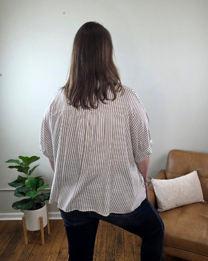 A woman with long brown hair smiles indoors beside a tan armchair and potted plant, wearing the Ryan Striped Short Sleeve Button Down Shirt and dark jeans, both from Cosmos and Clover Boutique in a light-colored room with wooden floors.