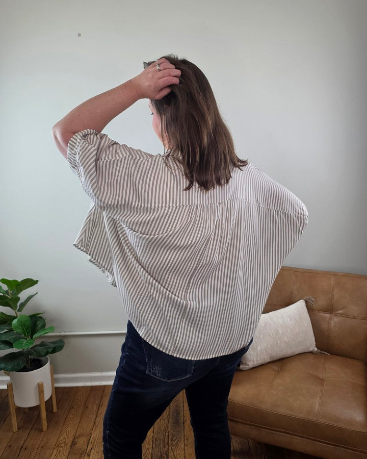 A woman with long brown hair smiles indoors beside a tan armchair and potted plant, wearing the Ryan Striped Short Sleeve Button Down Shirt and dark jeans, both from Cosmos and Clover Boutique in a light-colored room with wooden floors.