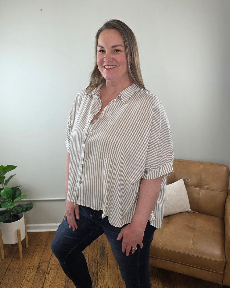 A woman with long brown hair smiles indoors beside a tan armchair and potted plant, wearing the ZENANA Ryan Striped Short Sleeve Button Down Shirt and dark jeans in a light-colored room with wooden floors.