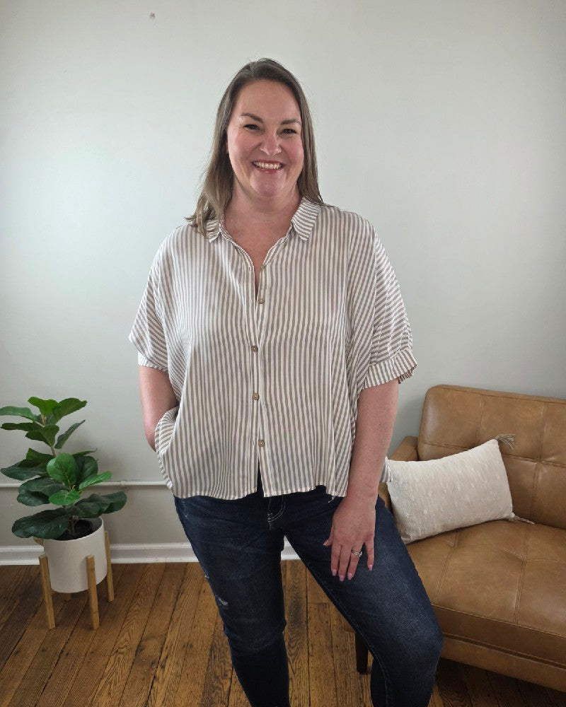 A woman smiles indoors wearing the ZENANA Ryan Striped Short Sleeve Button Down Shirt with dark jeans, one hand in her pocket. Behind her are a tan loveseat with a white cushion and a potted plant on the wooden floor.