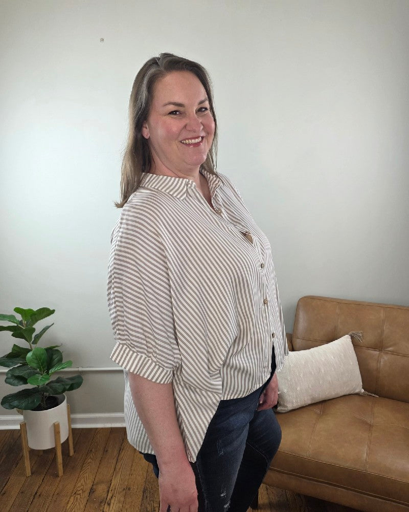 A woman with long brown hair smiles indoors beside a tan armchair and potted plant, wearing the Ryan Striped Short Sleeve Button Down Shirt and dark jeans, both from Cosmos and Clover Boutique in a light-colored room with wooden floors.
