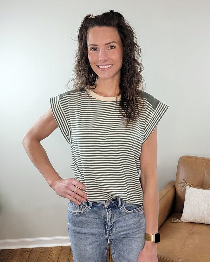 A woman with curly brown hair smiles indoors, modeling the Staccato Jules Striped Top and blue jeans. She wears a smartwatch, stands with one hand on her hip, and a tan couch with a pillow is in the background.
