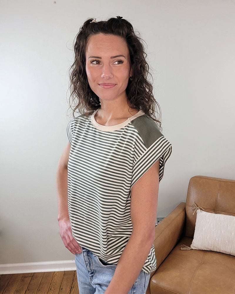 A woman with curly brown hair models the Staccato Jules Striped Top, a green and white short-sleeve style. She poses indoors with one hand in her jeans pocket, smiling softly beside a tan couch against a light wall.