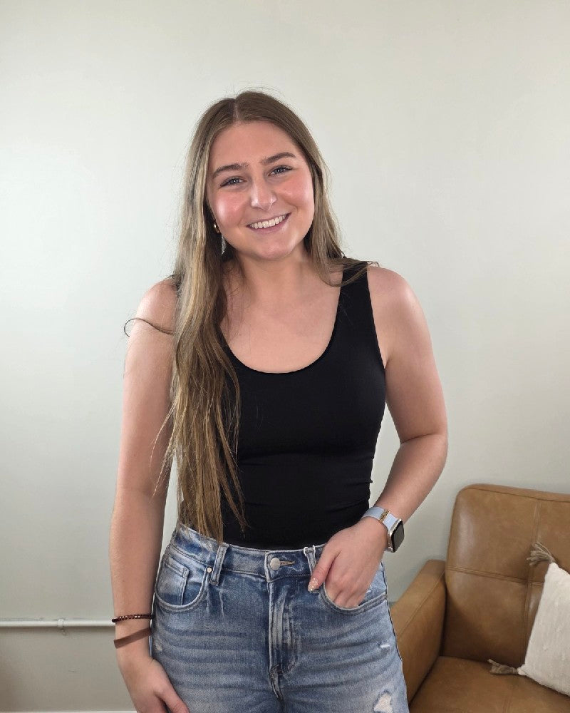 A young woman with long brown hair smiles indoors, wearing a ZENANA Seamless Tank Top with Built In Bra, blue jeans, and a smartwatch. A brown leather couch and a white pillow are visible in the background.