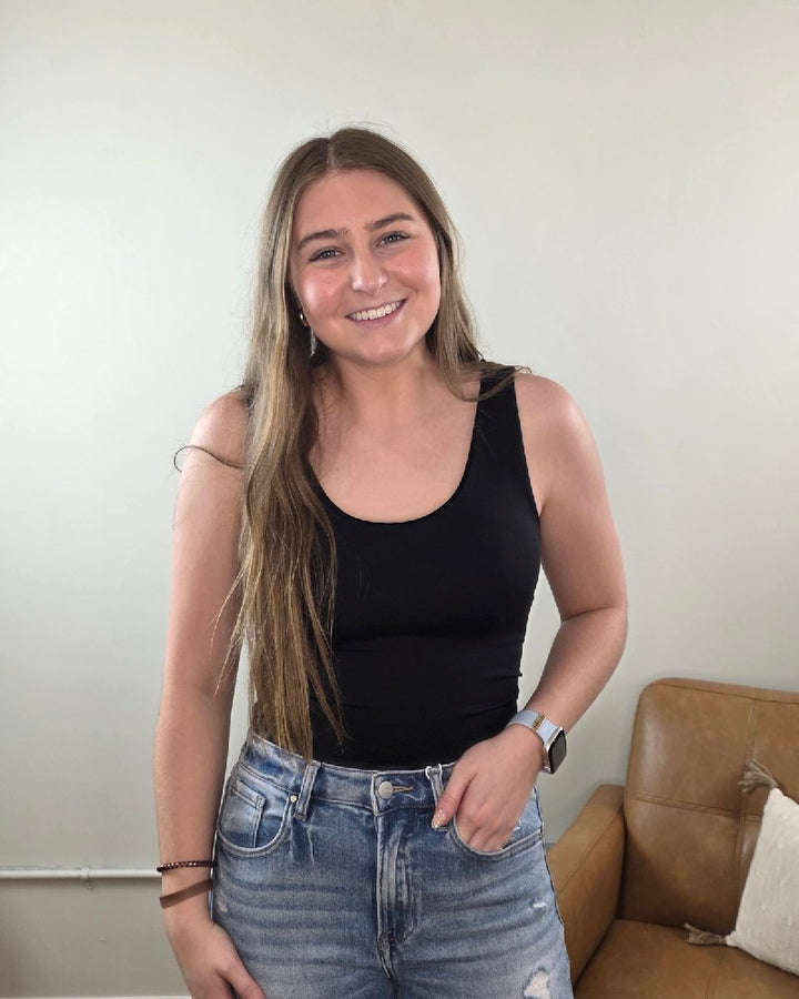 A young woman with long brown hair smiles indoors, wearing a ZENANA Seamless Tank Top with Built In Bra, blue jeans, and a smartwatch. A brown leather couch and a white pillow are visible in the background.