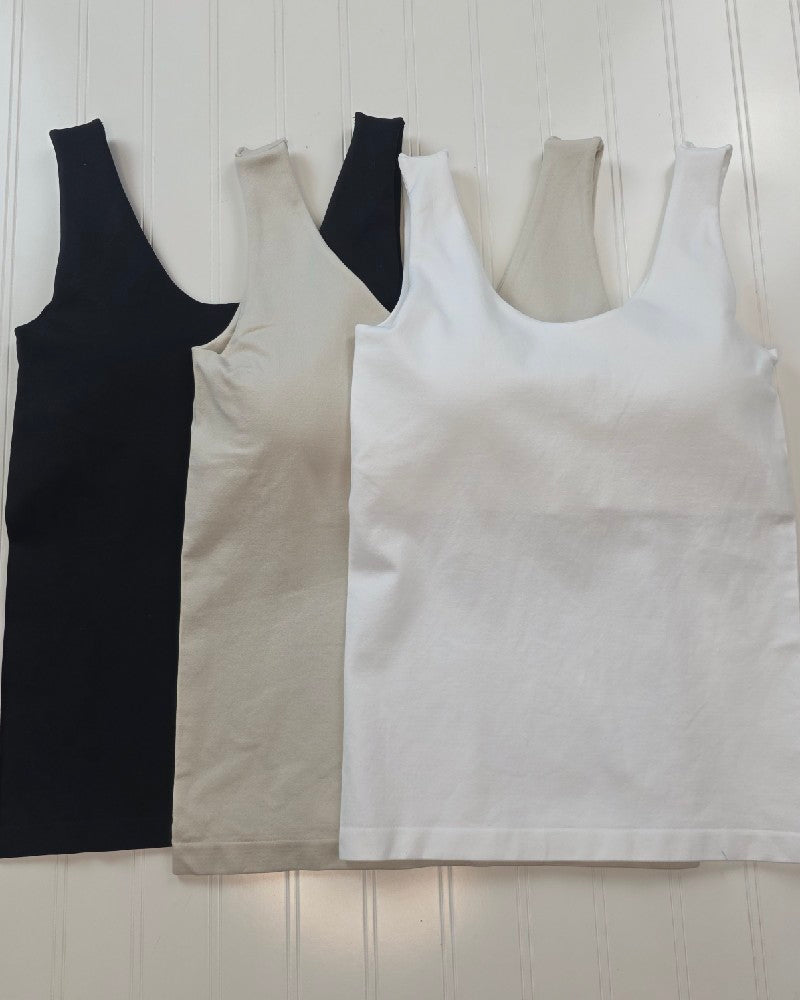 Three ZENANA Seamless Tank Tops with Built In Bra in black, beige, and white are arranged side by side on a textured white surface. Each offers comfort and support with its integrated bra design.
