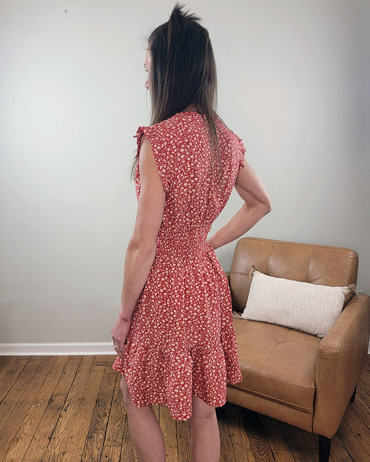 A woman stands indoors on a wooden floor, facing away from the camera, wearing the Staccato Savannah Dress—a red floral dress with ruffled sleeves, hem, and smocked waist. A tan armchair with a white pillow is in the background against a plain wall.