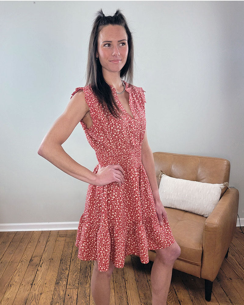 A woman with long brown hair wears a sleeveless red Staccato Savannah Dress with a smocked waist. She stands on a wooden floor beside a tan armchair with a white pillow against a plain light-colored wall.