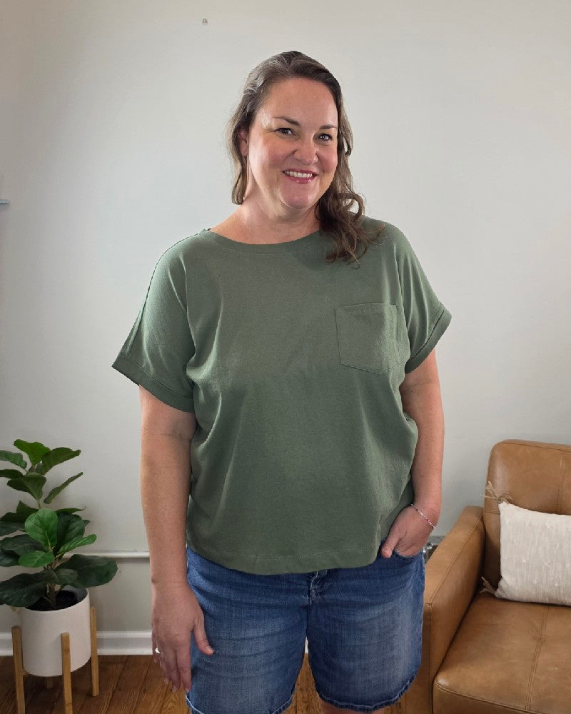 A woman with wavy brown hair, wearing the ZENANA Sibyl Cotton Tee with Front Pocket in green and denim shorts, stands smiling in a living room next to a plant and a tan sofa with a white pillow.