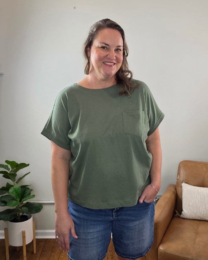 A woman with wavy brown hair, wearing the ZENANA Sibyl Cotton Tee with Front Pocket in green and denim shorts, stands smiling in a living room next to a plant and a tan sofa with a white pillow.