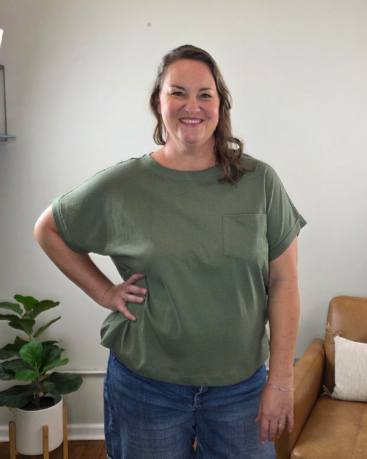 A woman with long brown hair stands smiling indoors with one hand on her hip, wearing the ZENANA Sibyl Cotton Tee with Front Pocket in green and a pair of jeans. A plant and brown armchair can be seen in the background.