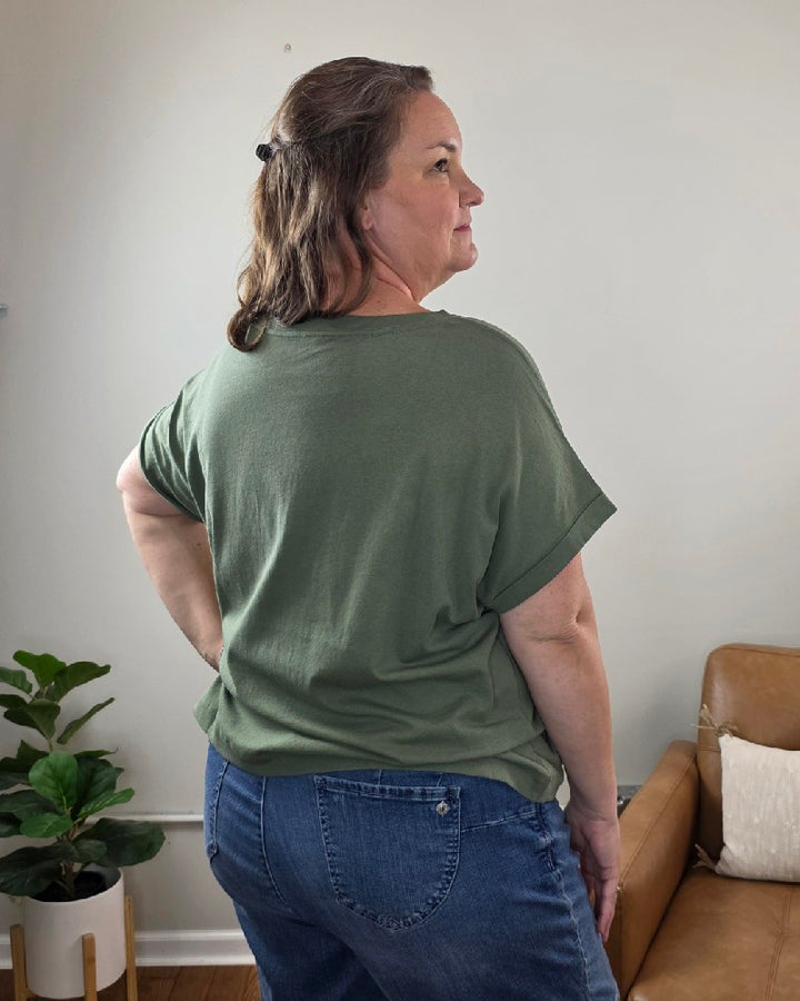 A woman with brown hair stands sideways in a room with light walls, a tan couch, and a potted plant. She is wearing the ZENANA Sibyl Cotton Tee with Front Pocket in green and blue jeans, looking over her shoulder with a neutral expression.