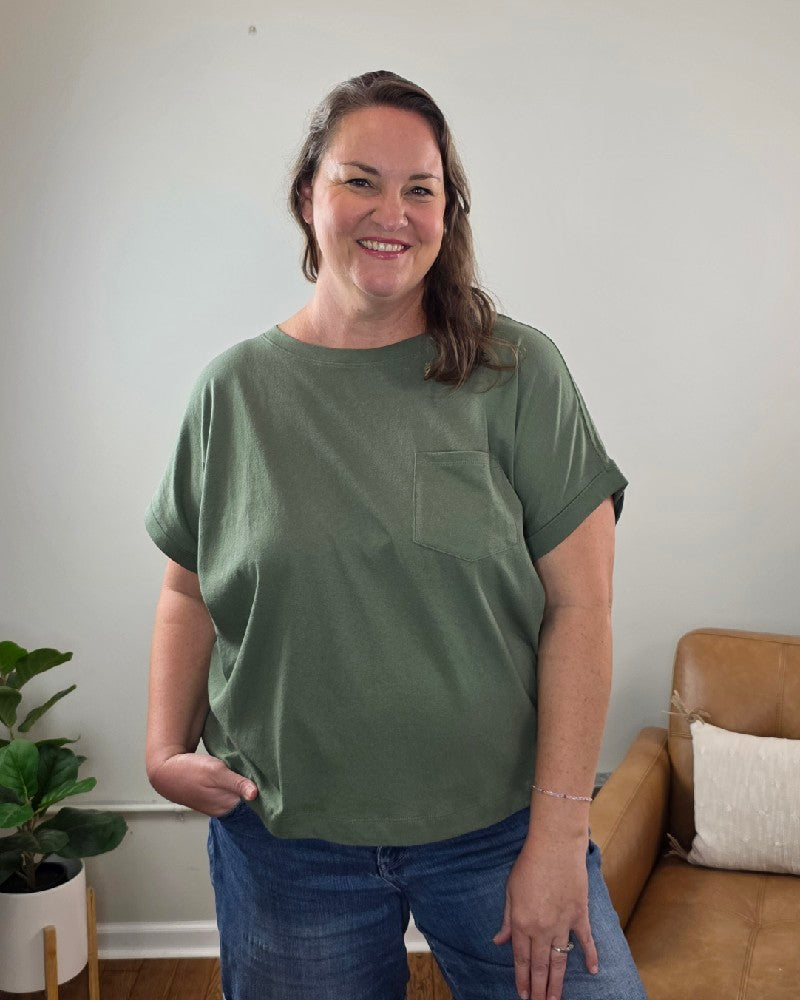 A woman stands indoors, smiling, wearing the ZENANA Sibyl Cotton Tee with Front Pocket in green and jeans. In the background, a tan couch and a potted plant are set against a light-colored wall.