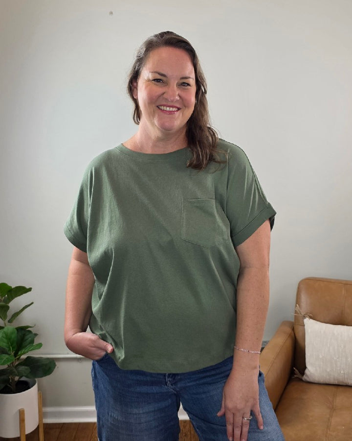 A woman stands indoors, smiling, wearing the ZENANA Sibyl Cotton Tee with Front Pocket in green and jeans. In the background, a tan couch and a potted plant are set against a light-colored wall.